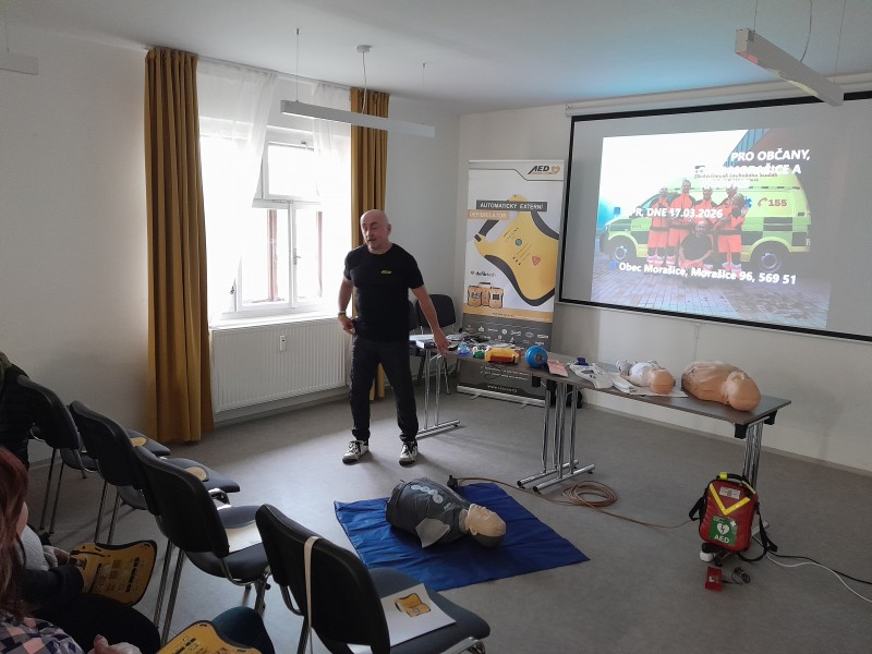 Na obrázku lektor předvádí a vysvětluje materiály a pomůcky pro první pomoc, které jsou rozložené na stole.